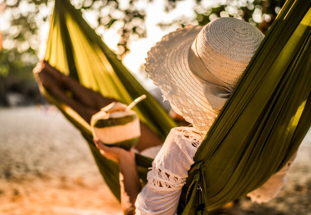 Frau, die sich in der Hängematte am Strand entspannt während sie einen Kokosnuss Drink trinkt.